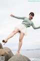 A woman in a green sweater and white skirt standing on rocks by the water.