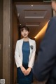 A woman in a blue dress and a white blazer standing in a hallway.